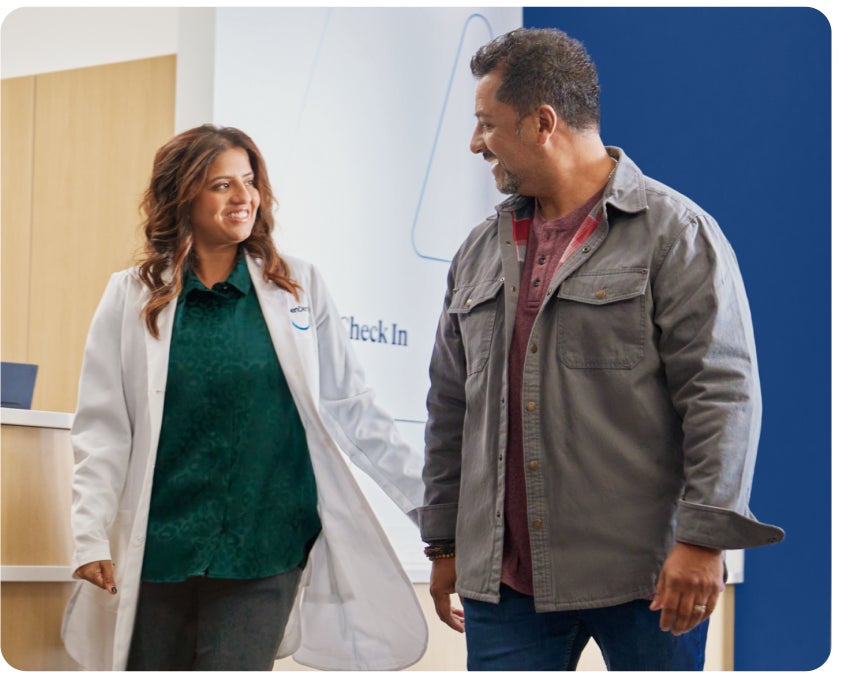 A person wearing a lab coat smiling and conversing with another person in casual attire, standing in a modern, professional setting.