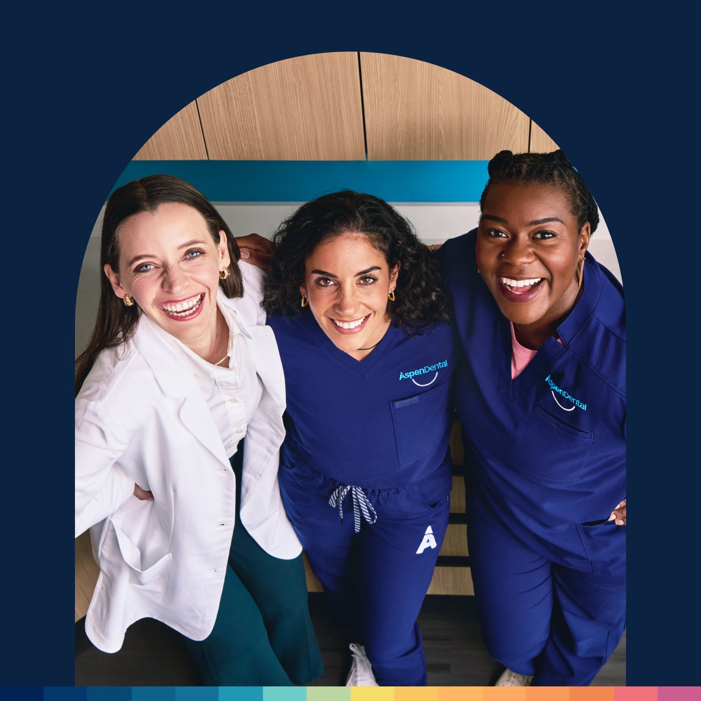 Three clear aligners experts, one in a white coat and two in blue scrubs, smile while standing together in a clinical setting.
