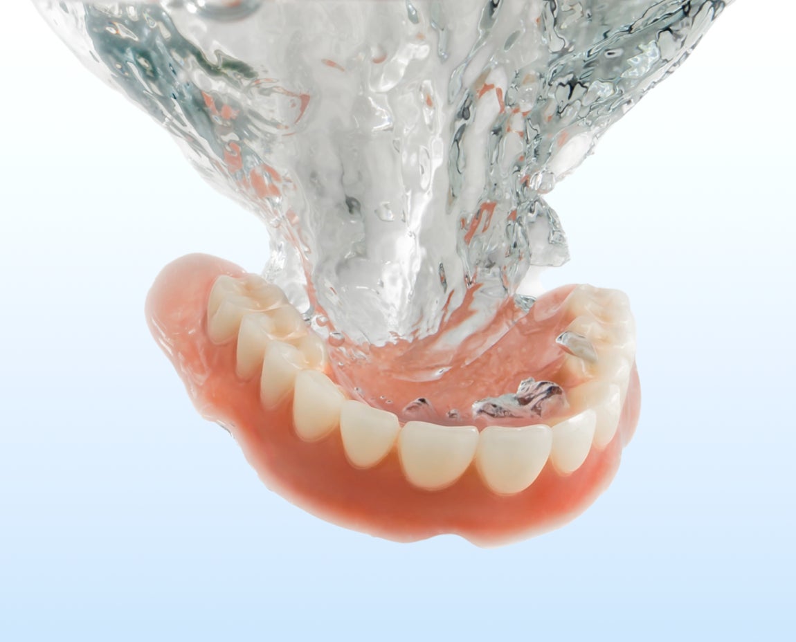 A set of lower dentures is submerged in clear water, against a light blue background, demonstrating the cleaning process.