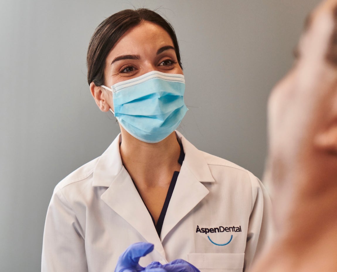 A dentist wearing a face mask and gloves, with Aspen Dental branding on her coat, speaking to a patient during a consultation about dental implant surgery.