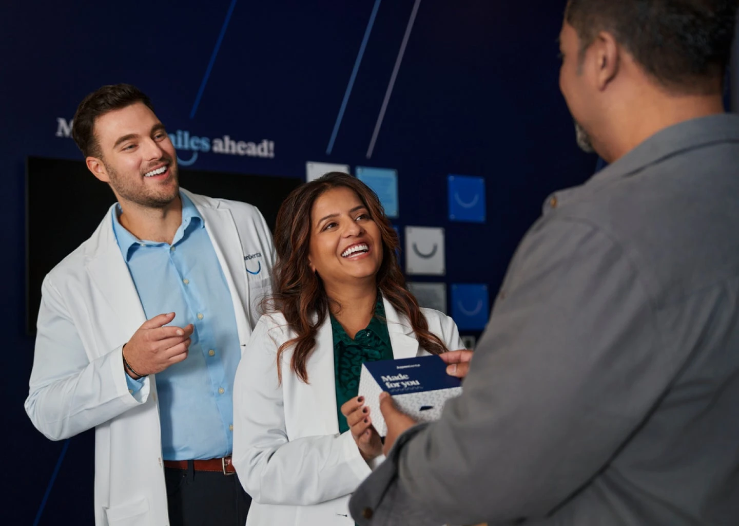 Two dental professionals in white coats smiling and talking to a person holding a product box in a dental office.