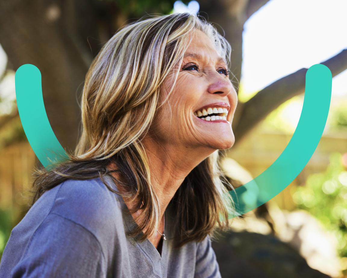 A joyful elderly woman with long hair, wearing a gray top, laughing outdoors with a blurred green background and teal graphics of a smile in the background.