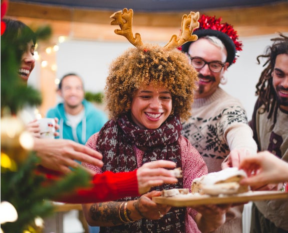 A group of friends at a festive holiday party, with a smiling woman wearing reindeer antlers holding a tray of cookies while others reach for treats in a joyful setting.
