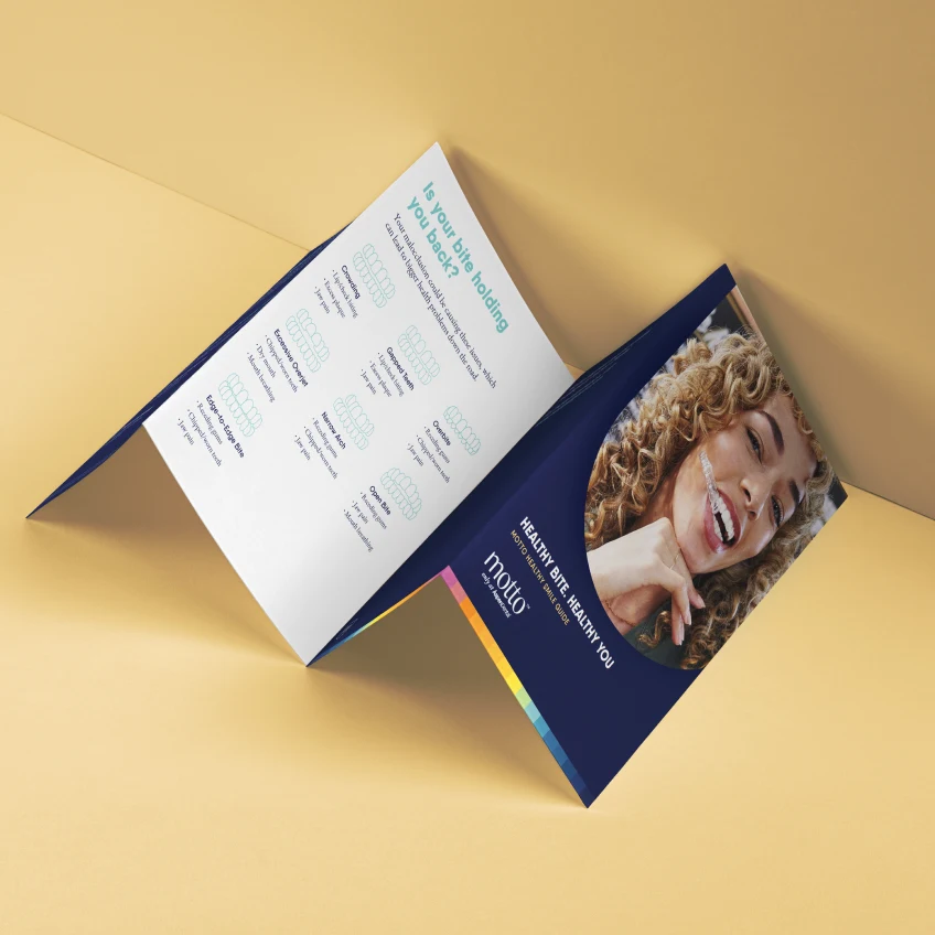 A woman holding clear aligners between her teeth on a pamphlet laid in a zig-zag way titled 'Healthy bite, healthy you. Motto healthy smile guide'