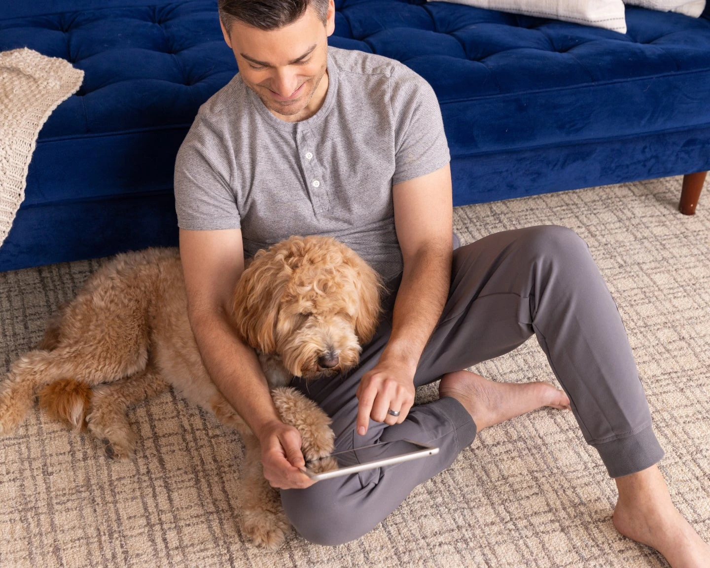 A man sitting with his dog on the floor, leaning against his blue couch working off his electronic device.