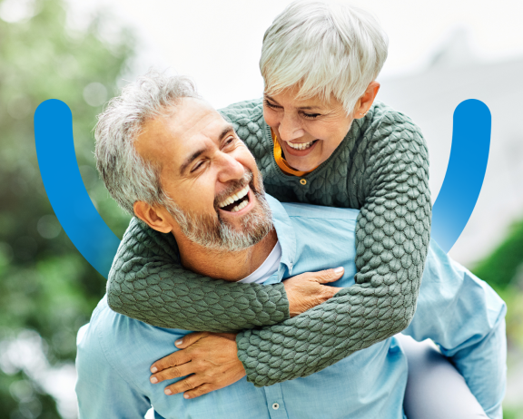 An older man giving a piggyback ride to an older woman outdoors, both laughing, with the Aspen Dental smile logo underlaid.