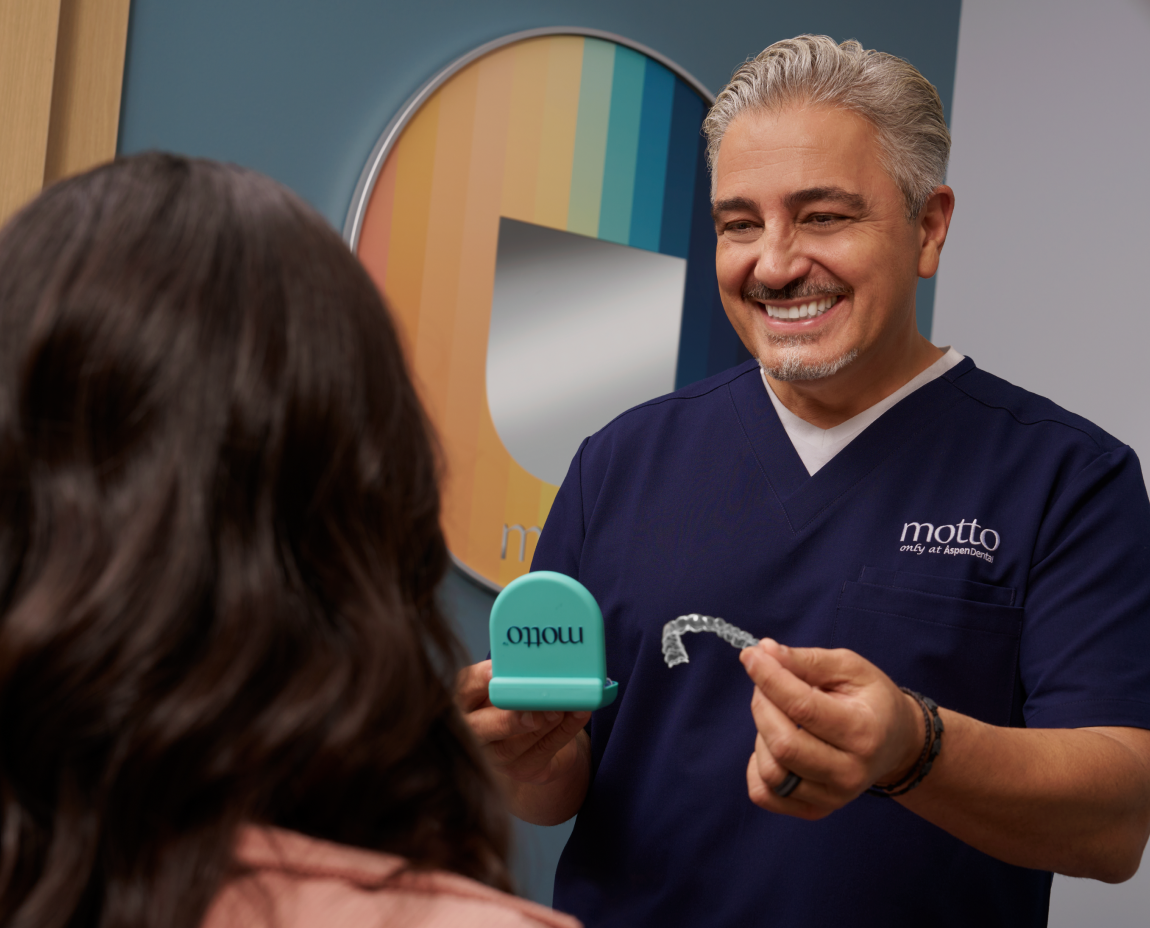 A Motto doctor smiling and holding clear aligners while he consults with a patient.