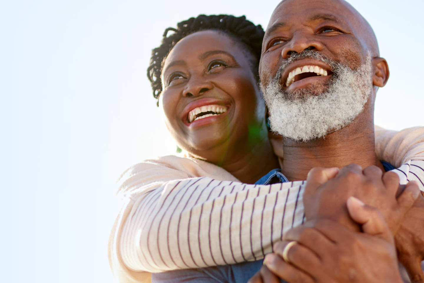 An older couple smiling joyfully, with the woman hugging the man from behind against a clear sky background.