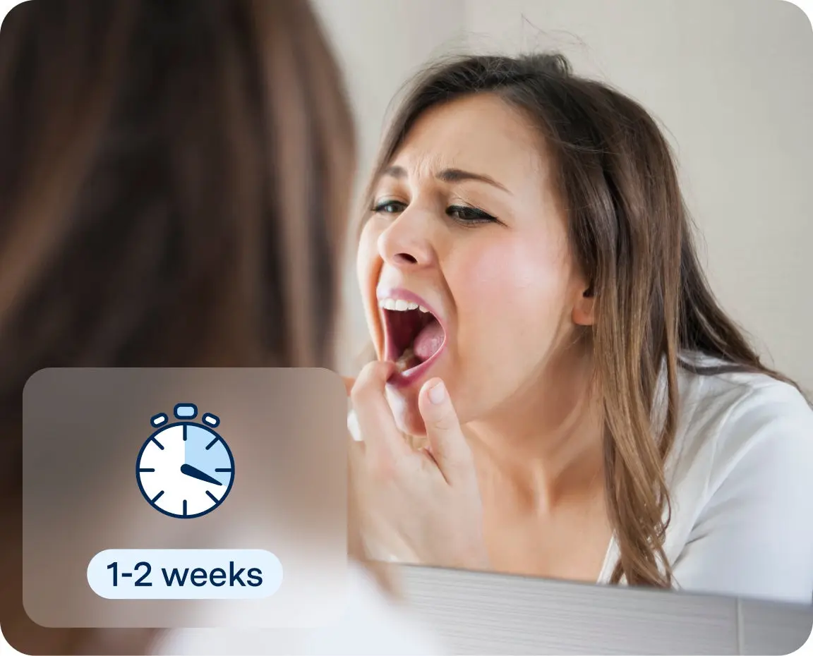 A woman uses a mirror to examine a sore spot on her tongue, a typical action when assessing healing time of canker sores and other oral health issues.