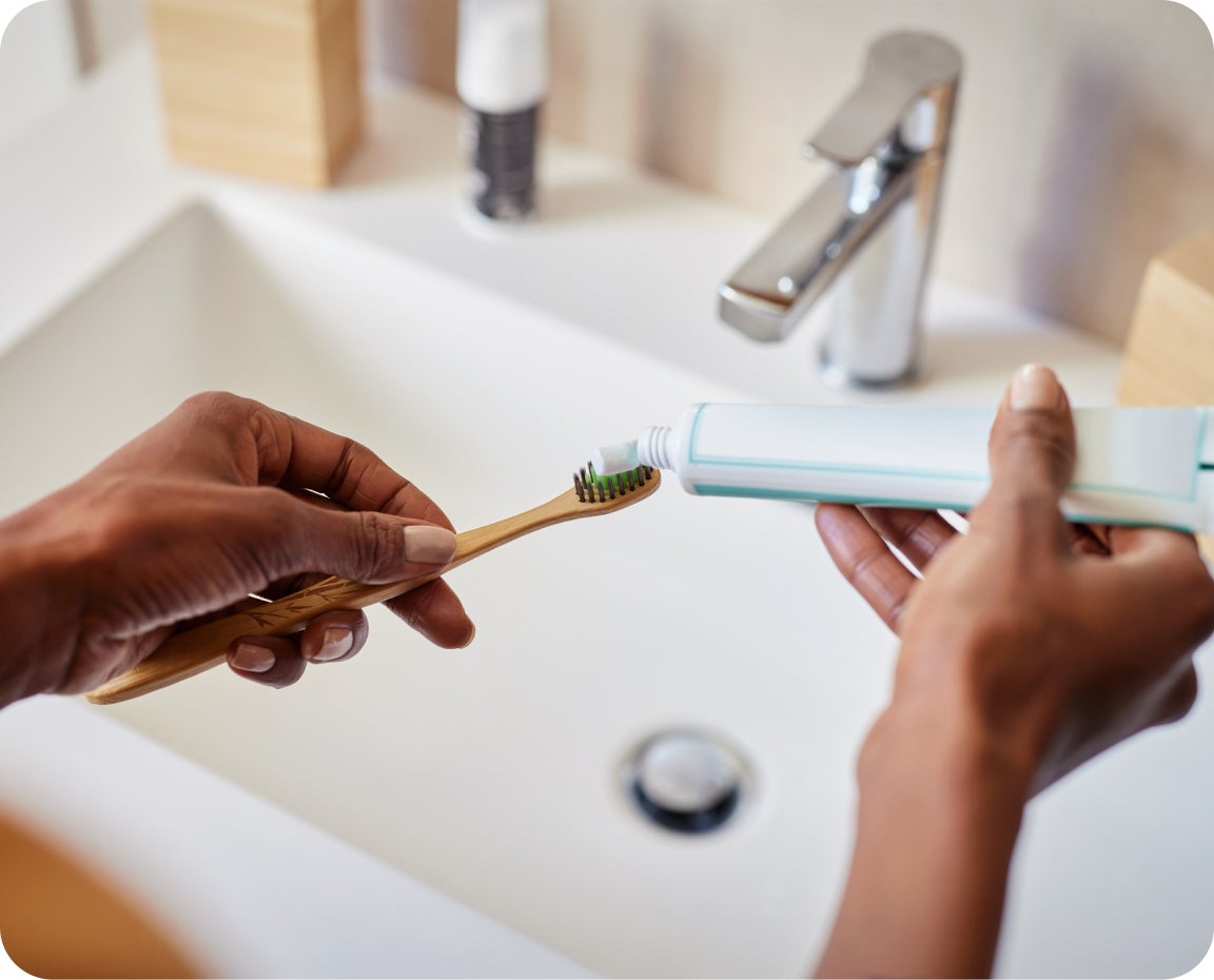  A person applies toothpaste to a bamboo toothbrush over a bathroom sink, practicing good oral hygiene to help reduce bad breath.