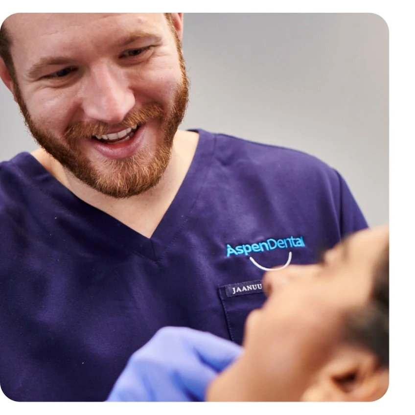 A bearded man in a navy blue shirt with "Aspen Dental" logo, smiles while interacting with a person lying down, partially visible in the foreground, possibly preparing for an emergency tooth extraction.
