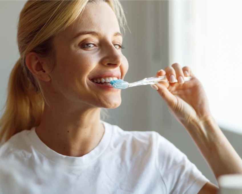 A woman with blonde hair in a ponytail is brushing her teeth with a blue toothbrush, smiling, and wearing a white shirt.