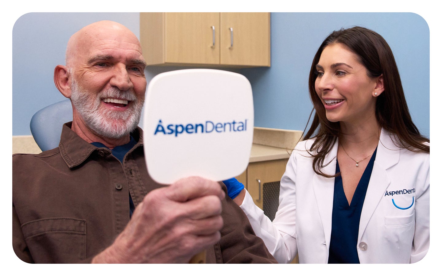 A happy Aspen Dental patient admires his smile in a handheld mirror after receiving dental implants. A dentist in a white coat smiles beside him, highlighting the confidence and benefits of tooth replacement solutions.
