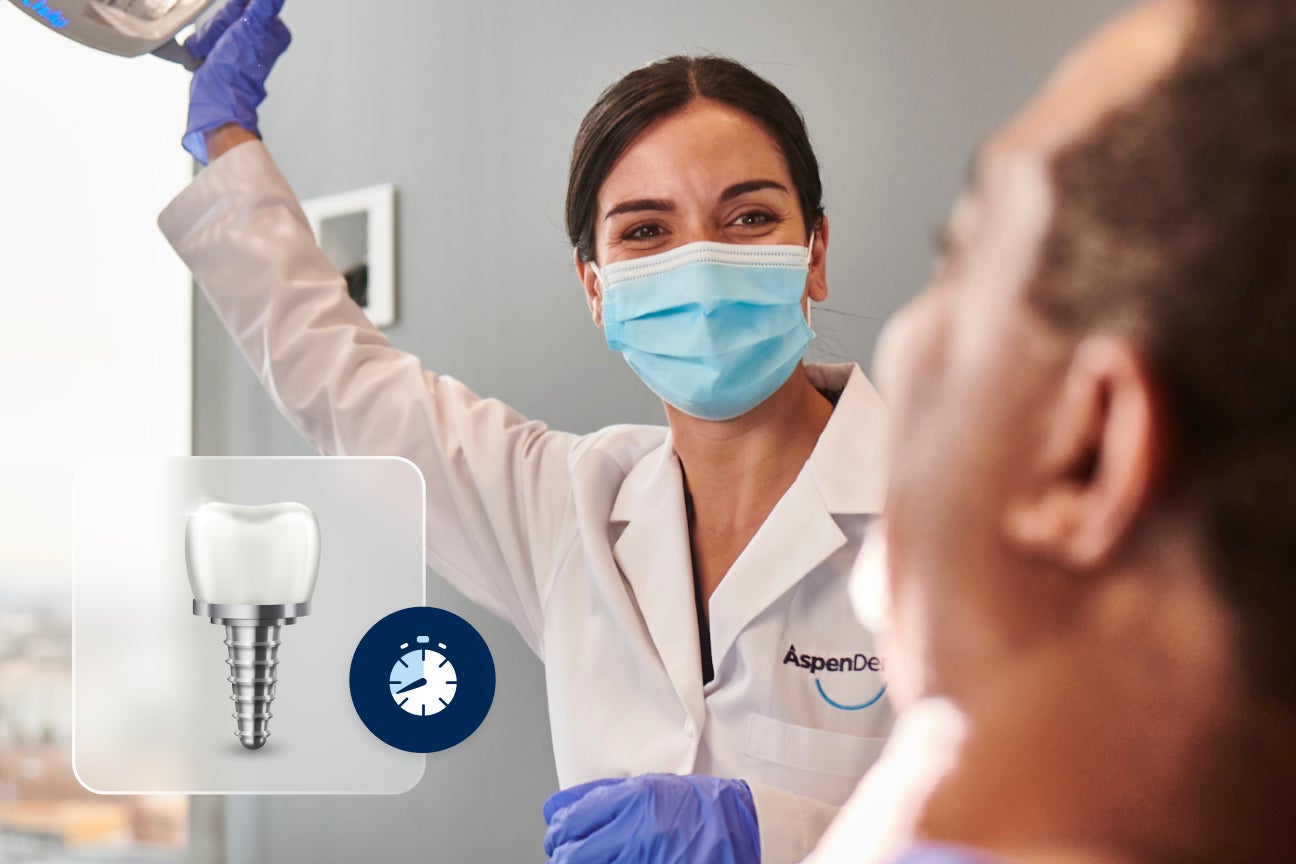 Image of an Aspen Dental dentist with a blue face mask over her mouth and nose, wearing blue gloves and a white coat. She is facing a dental light over her smiling patient who is blurred out. In the bottom left corner there is a small graphic of a single dental implant with a screw at the bottom and a illustration of a clock to the right of that.