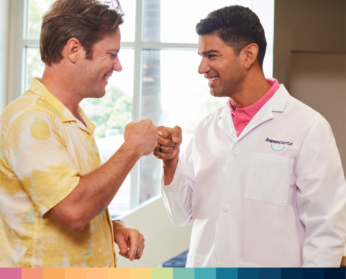 An Aspen Dental dentist wearing a white coat and a young male patient greeting each other with a fist bump.