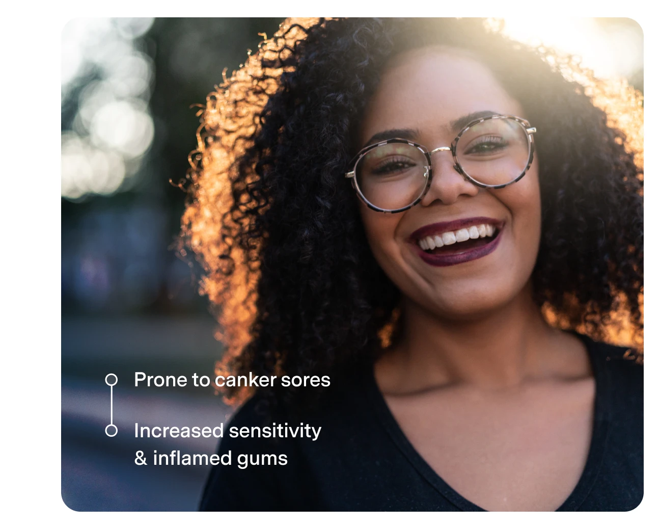 A smiling woman with curly hair and glasses, standing outdoors. Text reads "Prone to canker sores" and "Increased sensitivity & inflamed gums" on the left side.