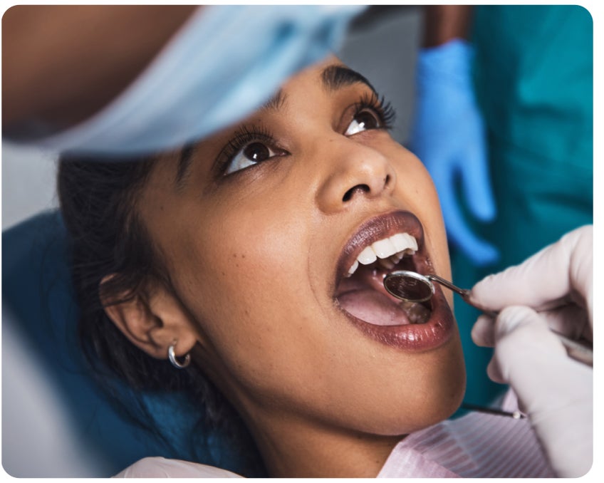 A person reclines in a dentist's chair with their mouth open while a dentist uses a mirror to examine their teeth.