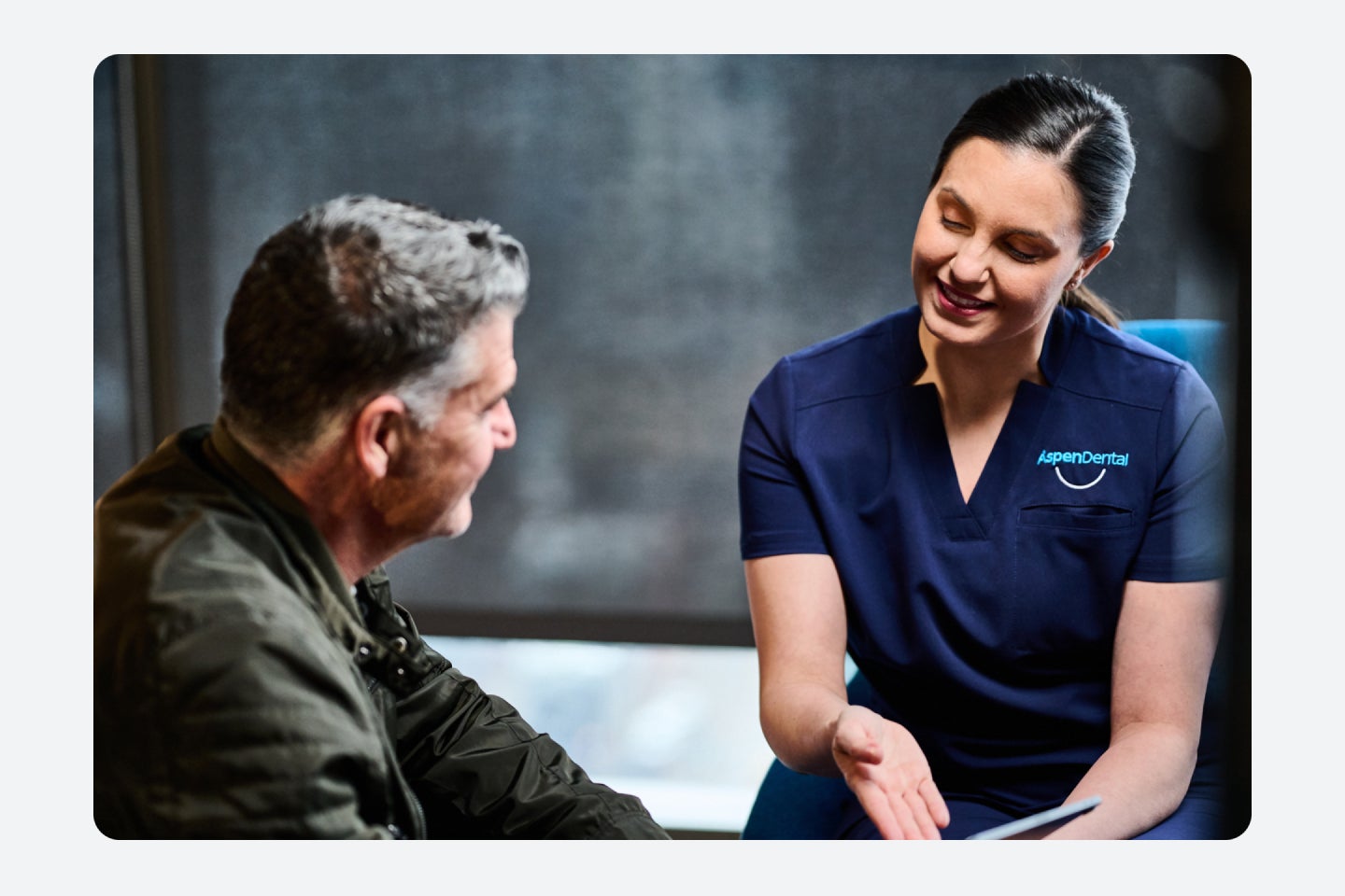 An Aspen Dental care team member, with dark black hair and dark navy scrubs is showing her patient, his dental appointment results.