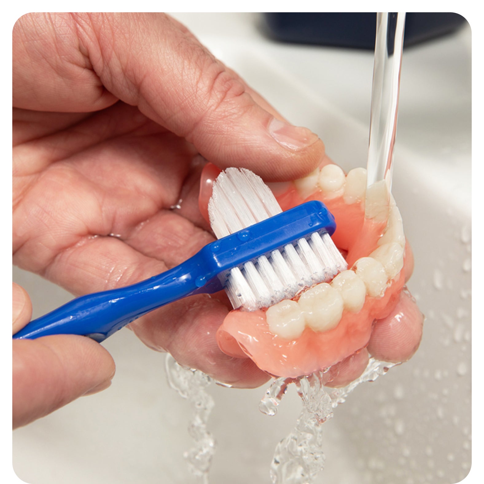  A close-up of hands cleaning dentures with a blue toothbrush under running water, emphasizing proper denture care for Aspen Dental.