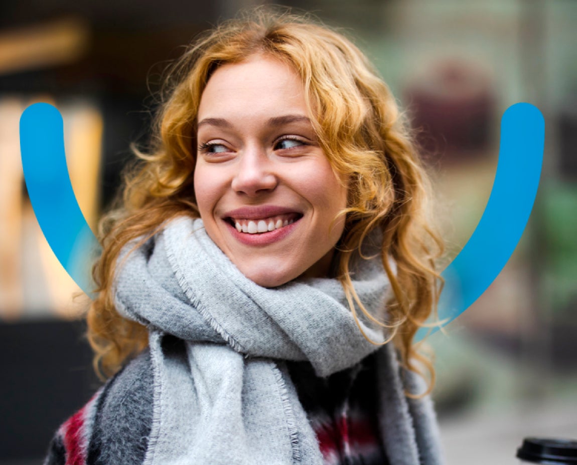 A woman with curly blonde hair is smiling and looking to the side while wearing a gray scarf and a checkered coat. The background is blurred and contains blue curved lines.