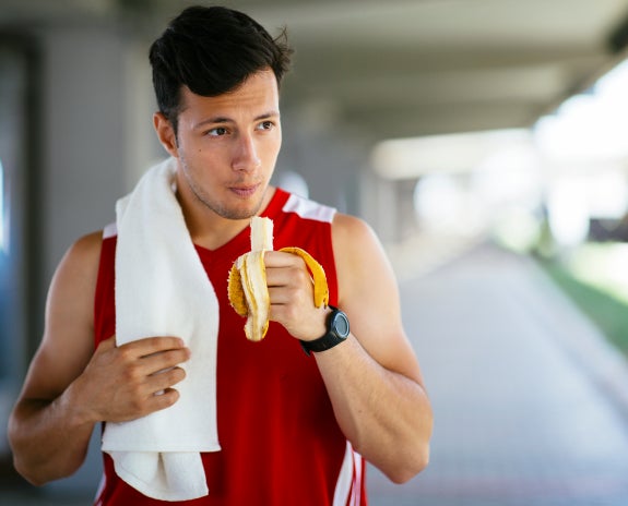 A young man in a red sleeveless shirt eating a banana while holding a white towel, appearing to have finished a workout.