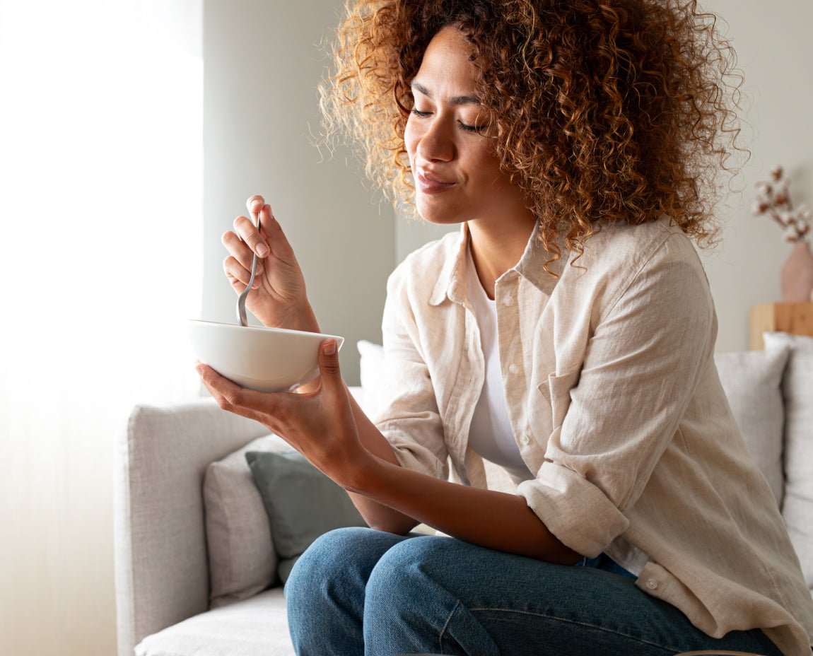 A woman sitting on a couch, smiling and enjoying a bowl of food, representing improved chewing efficiency and quality of life after dental implant surgery at Aspen Dental.