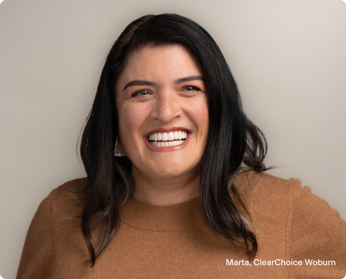 A woman with dark hair wearing a brown sweater smiles warmly while posing in front of a neutral beige background. The image conveys a sense of happiness and confidence.