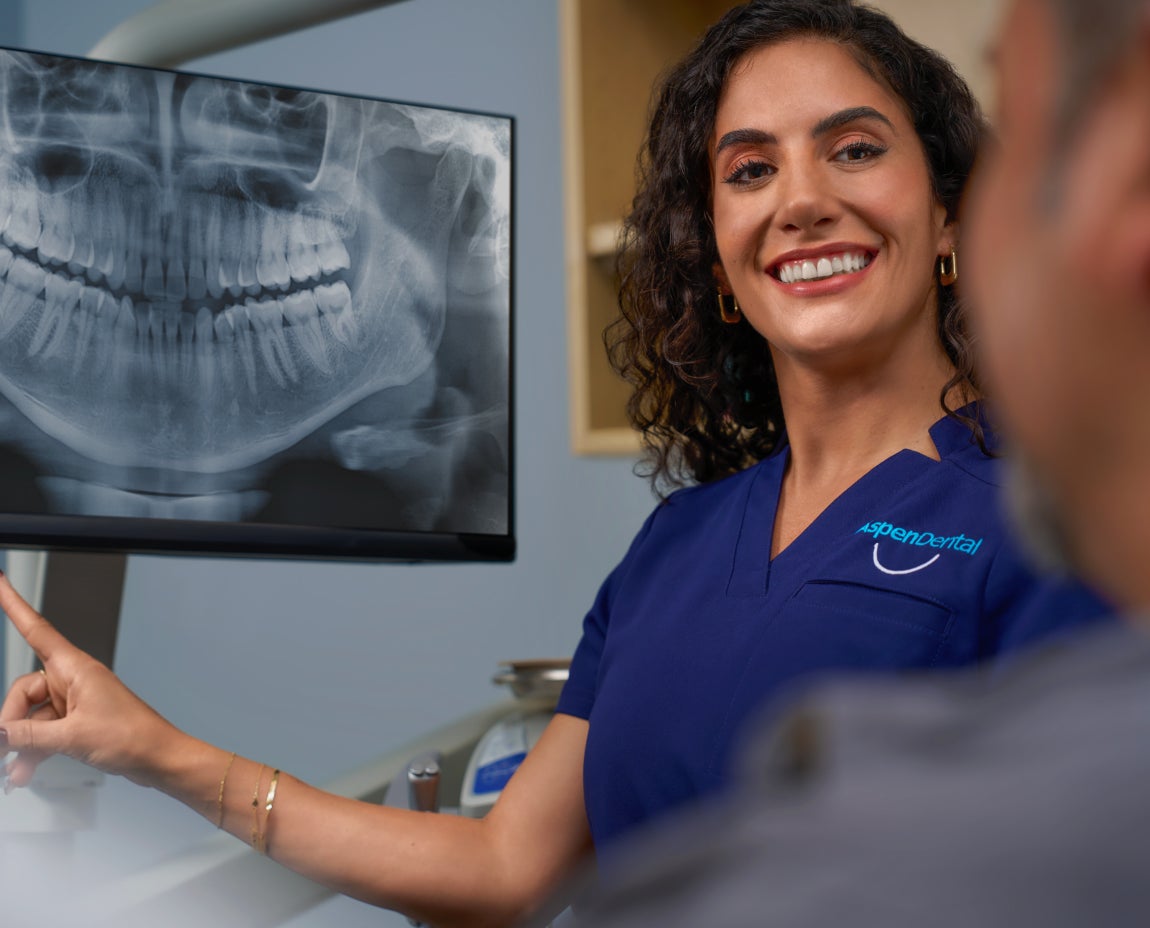 A woman in a blue scrub with an Aspen Dental logo is pointing at an x-ray to a client.