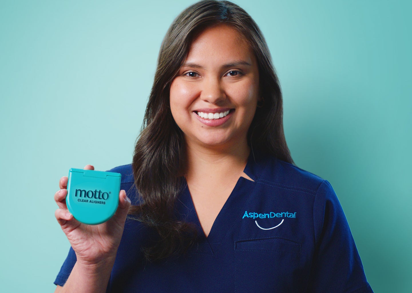 A smiling female dental assistant in an Aspen Dental uniform holding a turquoise Motto aligners case against a teal background.