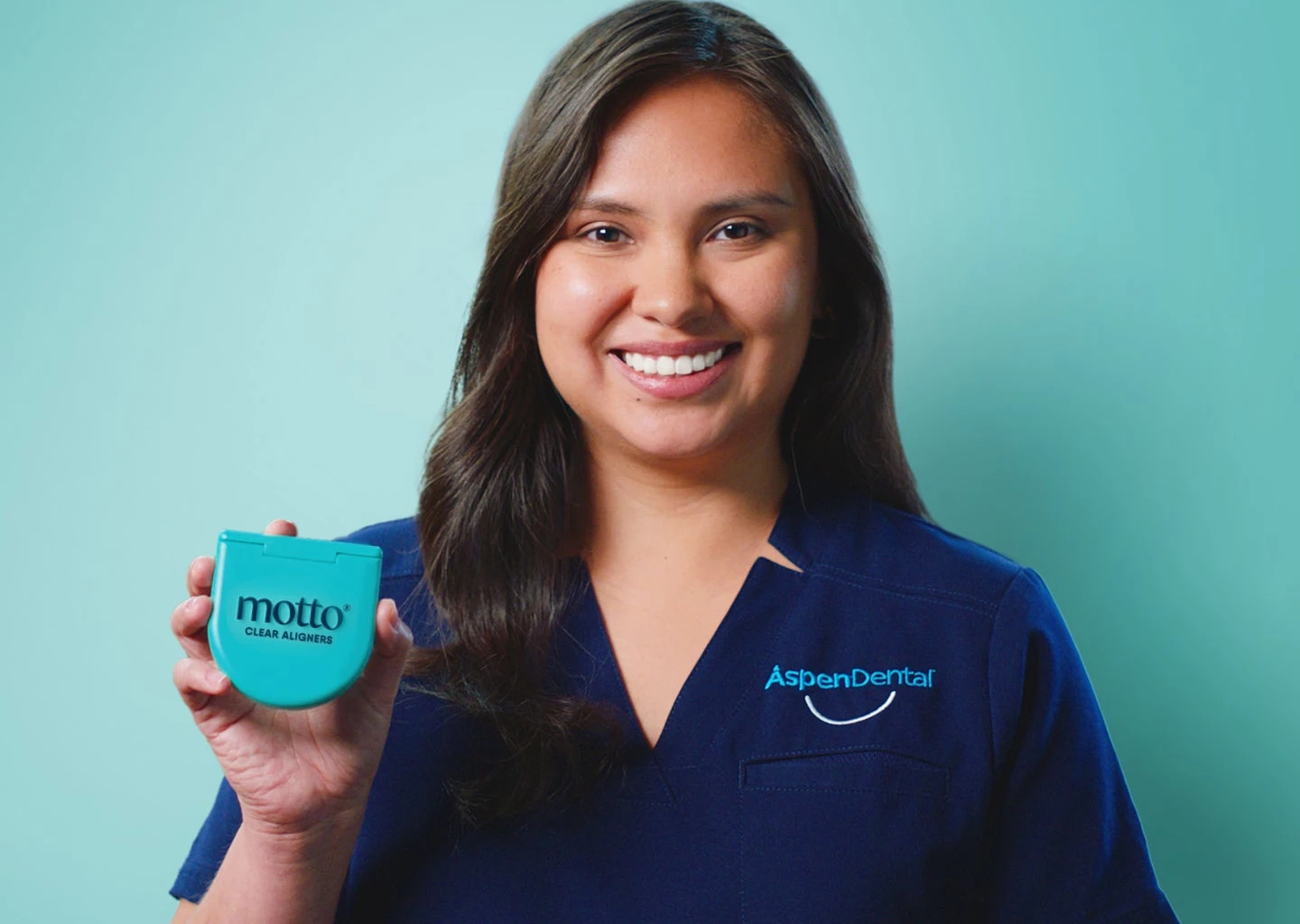 A smiling female dental assistant in an Aspen Dental uniform holding a turquoise Motto aligners case against a teal background.