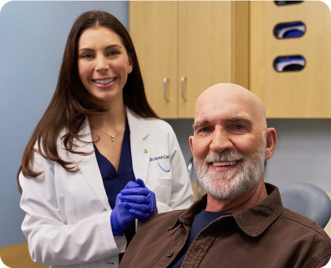 A dentist in a white coat stands beside a smiling older male patient seated in a dental chair after treatment.