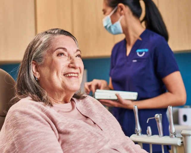 An older woman smiling in a dental clinic during her in-office periodontal disease treatment, while a dental assistant works in the background.