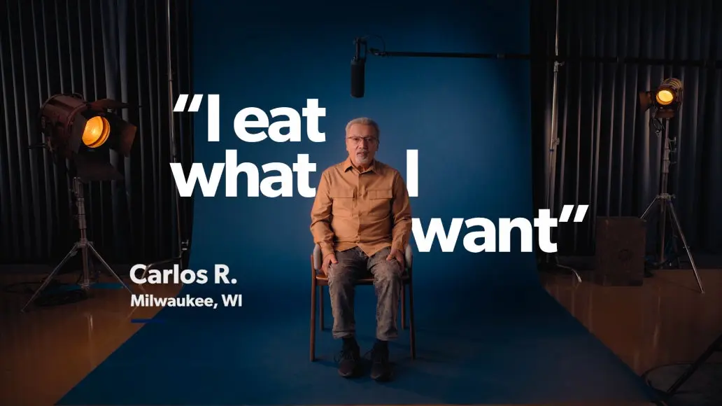 A man sitting on a stool in front of a blue backdrop with the text "I eat what I want." The name "Carlos R." and location "Milwaukee, WI" are displayed on the left. Studio lights and microphone are visible.