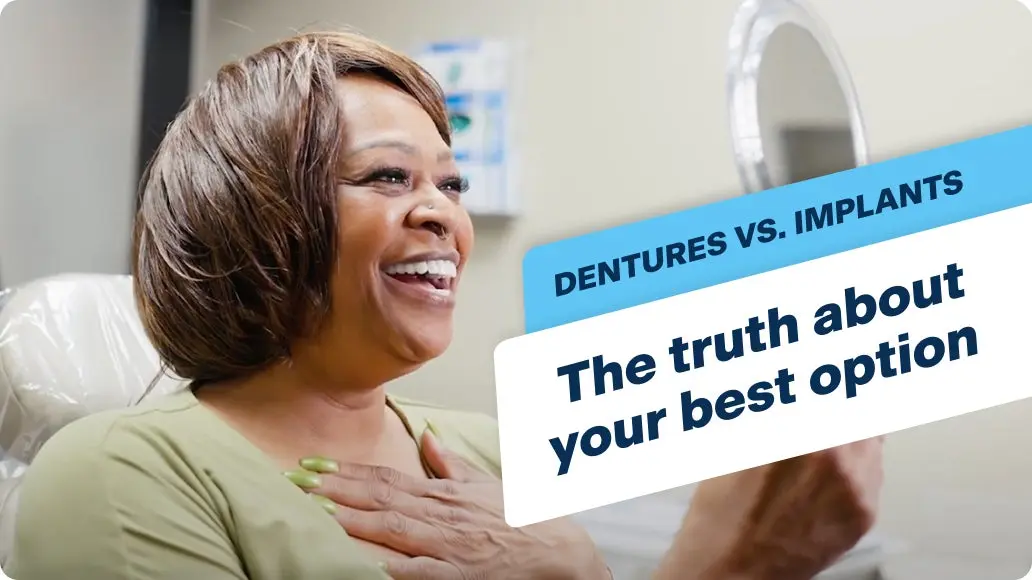A smiling woman with shoulder-length brown hair wears a light green top while seated in a dental office. She smiles into a hand held mirror as she admires her new dental implants, with text saying "Dentures vs implants, the truth about your best option".
