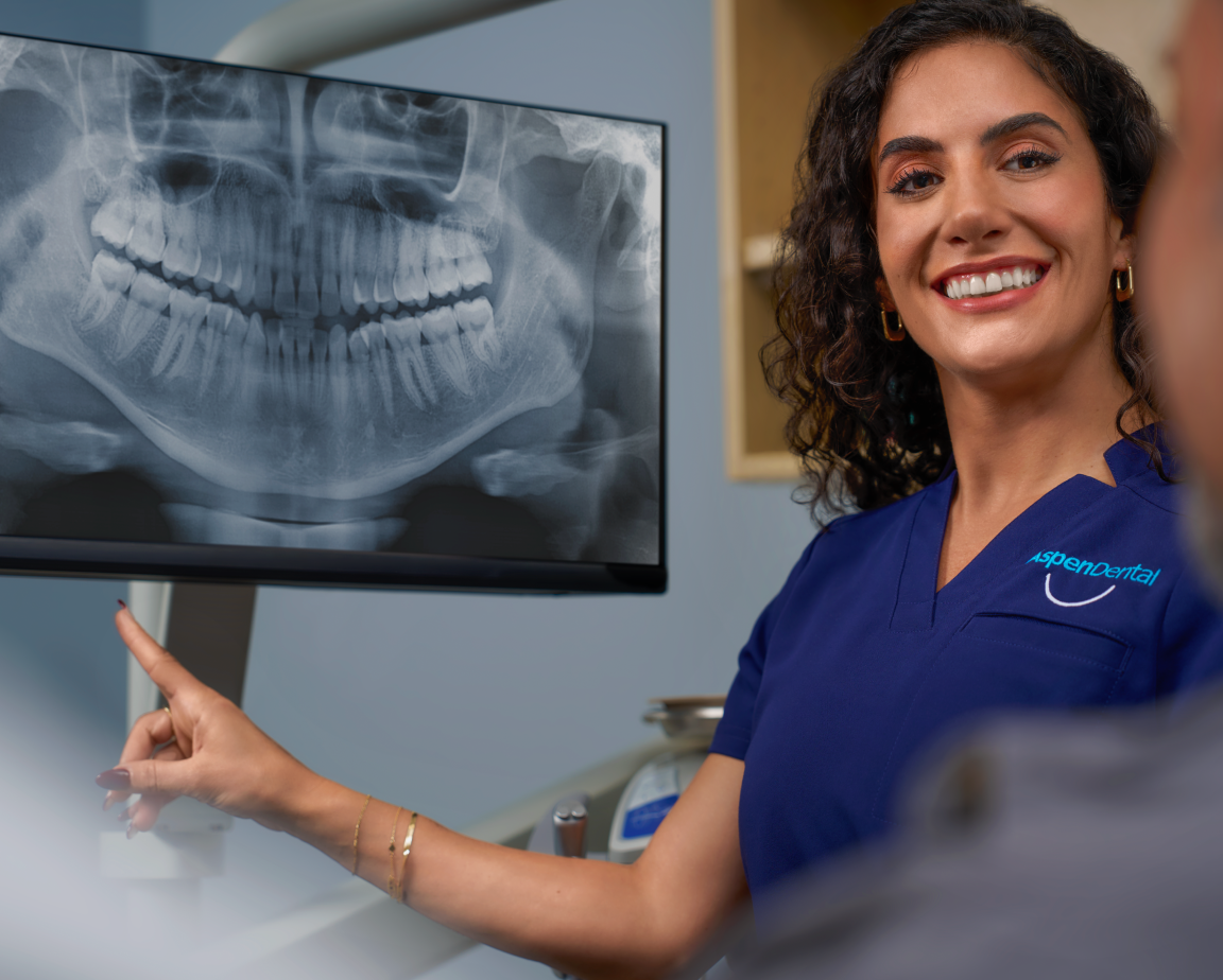 A female dentist in blue scrubs smiles and points to a dental X-ray displayed on a monitor in an office.