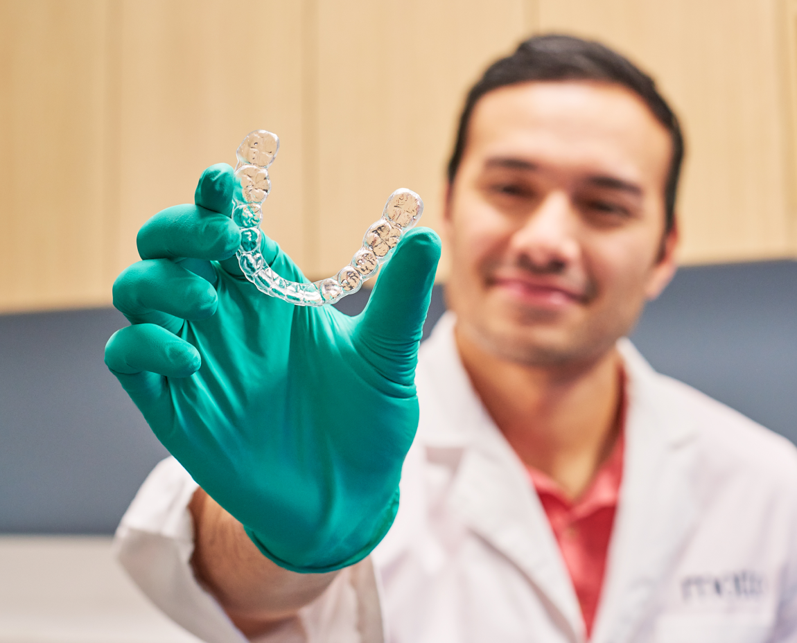 A person wearing a white lab coat and green gloves holds a clear aligner, smiling in the background.