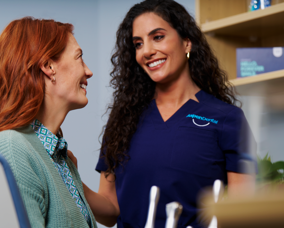 Red-haired patient smiling at her dentist, a woman with curly hair dressed in blue scrubs, in a dental clinic, who is also smiling.