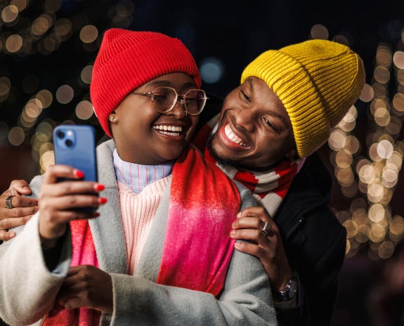 A happy couple wearing colorful winter hats and scarves taking a selfie together outdoors at night, with festive holiday lights glowing in the background.
