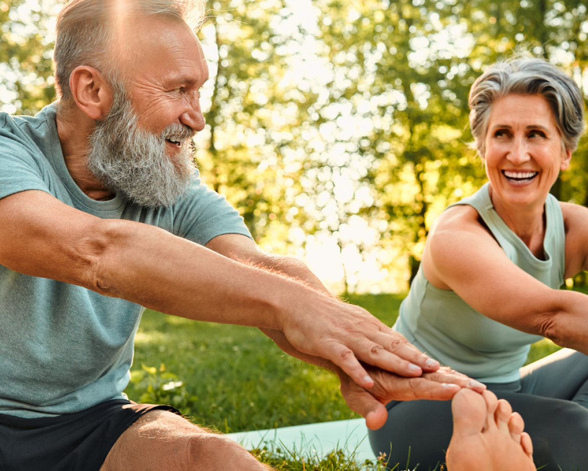 A smiling older man and woman sitting and leaning forward while stretching their arms and touching their foot during an outdoor exercise session in a park on a sunny day.