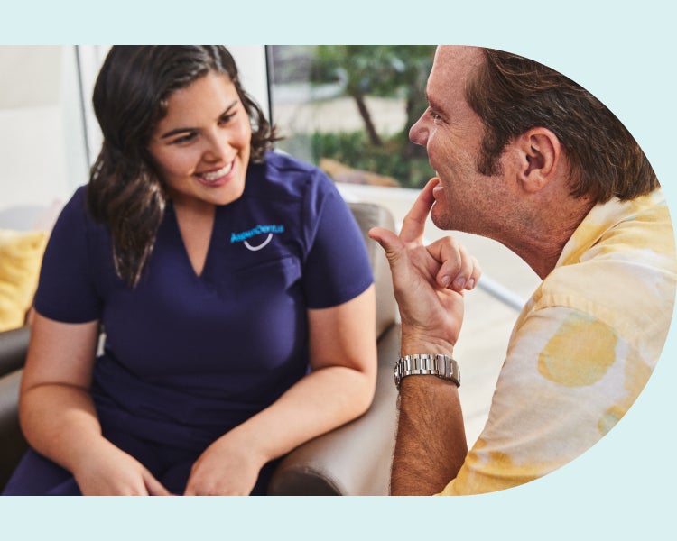 A man and a woman in scrubs sit in a dental office. The man points to his teeth while talking to the woman.