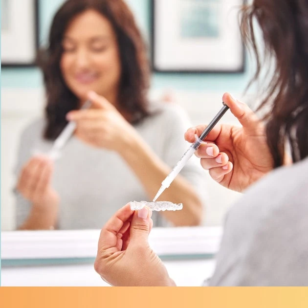 A person is holding a dental aligner in one hand and a syringe in the other, while standing in front of a mirror.