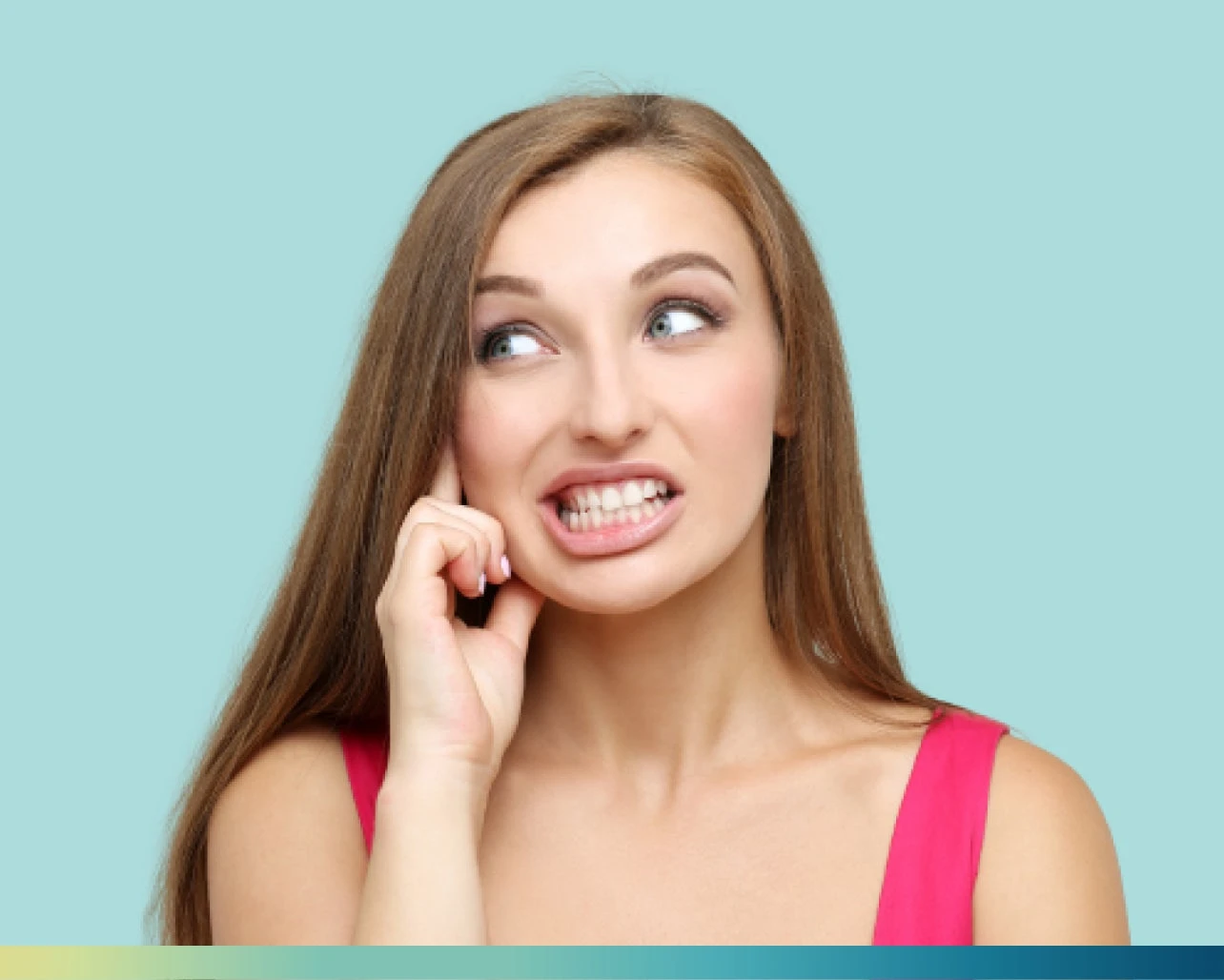 A woman with long brown hair and wearing a pink top has a slightly tense expression, clenching her teeth while awake, and touching her face. The background is a solid light blue.