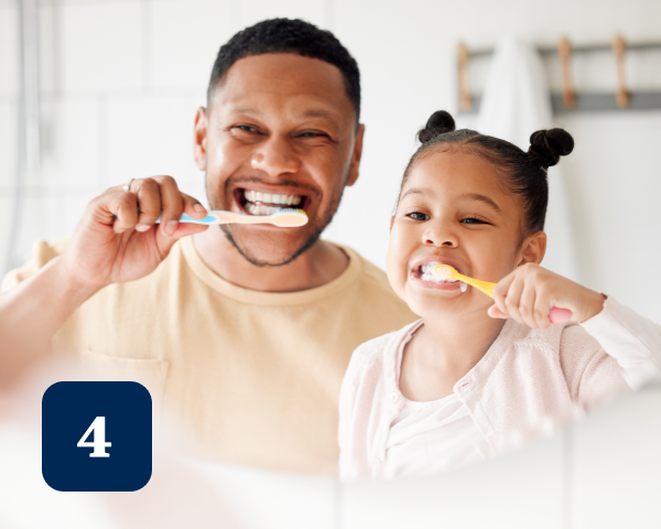 A man and a young girl are smiling and brushing their teeth together in a brightly lit bathroom.