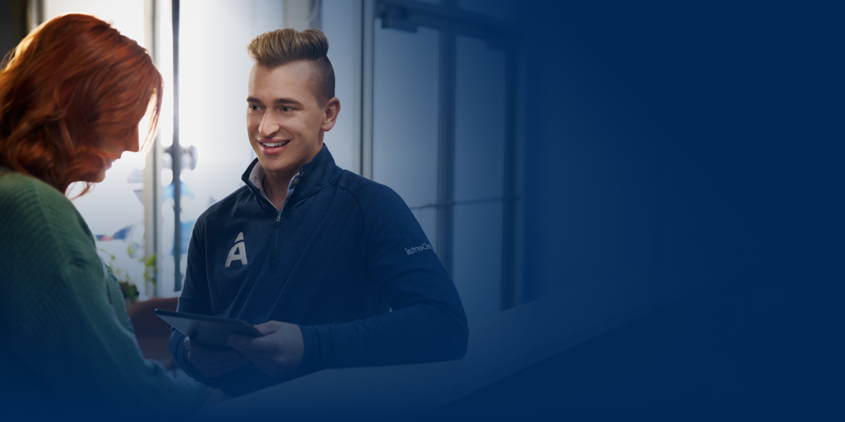 A woman with red hair is engaging in a conversation with a dental professional holding a tablet, wearing a navy jacket with a large letter "A" on it. The background is blurred.