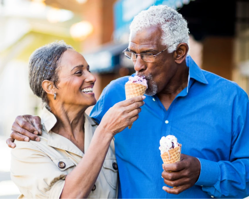 Senior couple enjoying ice cream together, with woman feeding man a bite, in casual outdoor setting.