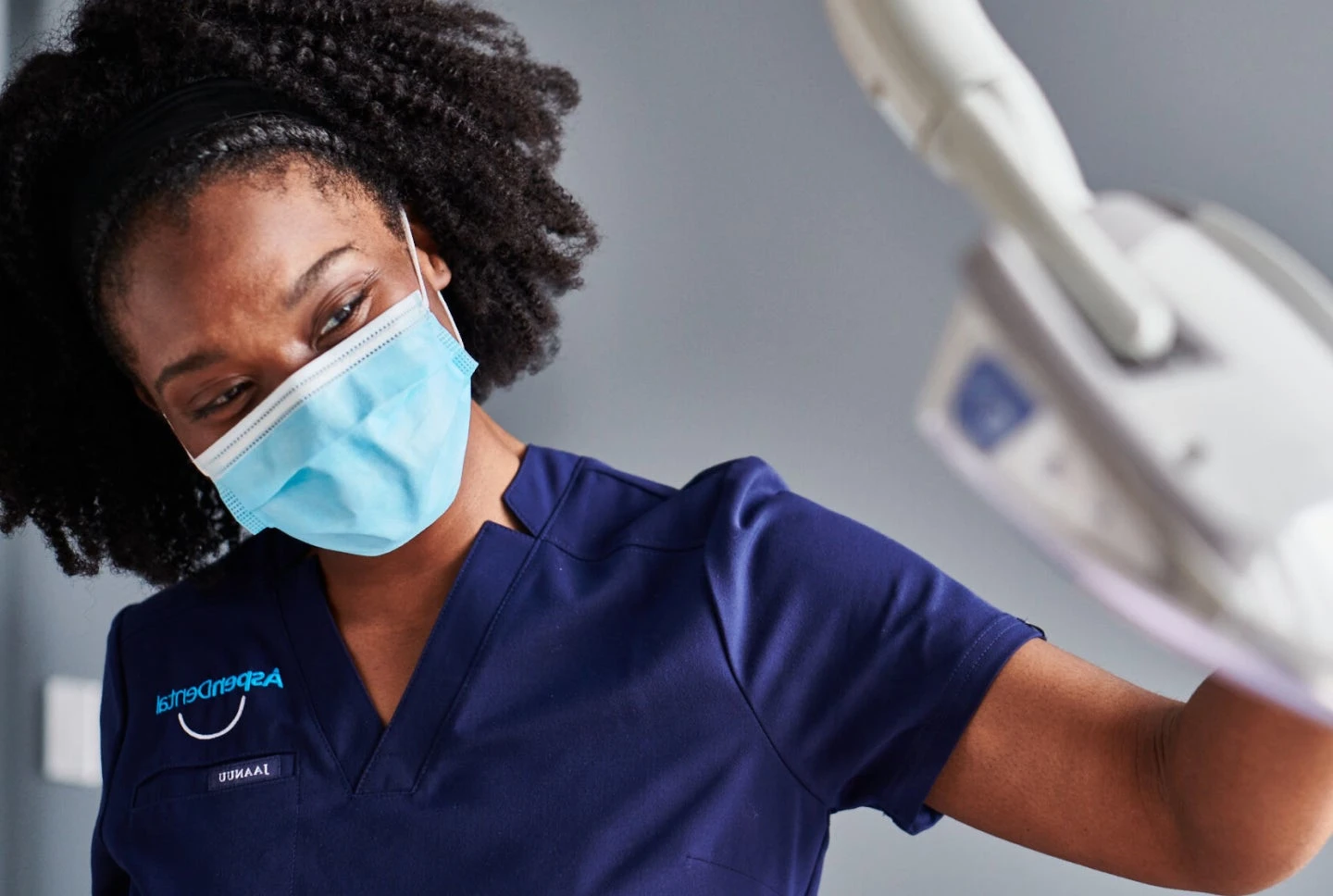 Aspen Dental professional wearing a mask and blue uniform, preparing an X-ray machine for a patient in a modern dental clinic.