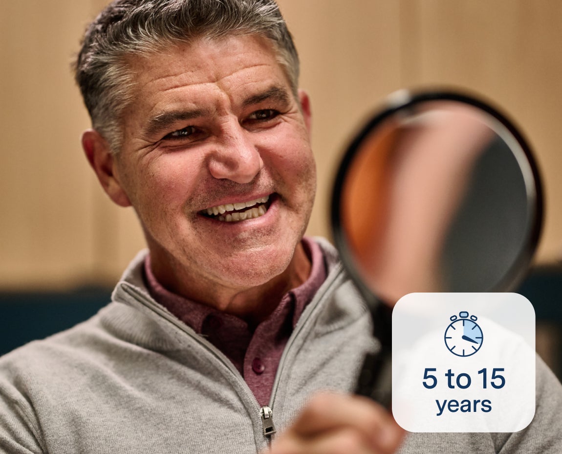 A smiling man holding a mirror to examine his teeth, symbolizing the durability of dental implants provided by Aspen Dental, which can last 5 to 15 years with proper care.