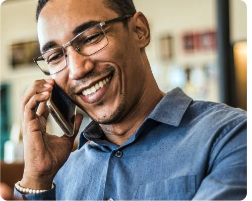 A man wearing glasses and a blue shirt is smiling while talking on a smartphone.