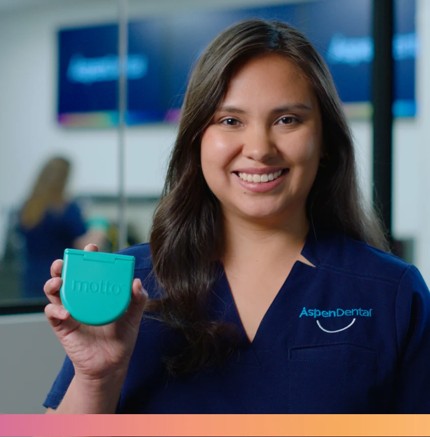 A woman wearing a navy blue Aspen Dental scrubs smiles while holding a turquoise dental product labeled "motto" in a dental office setting.