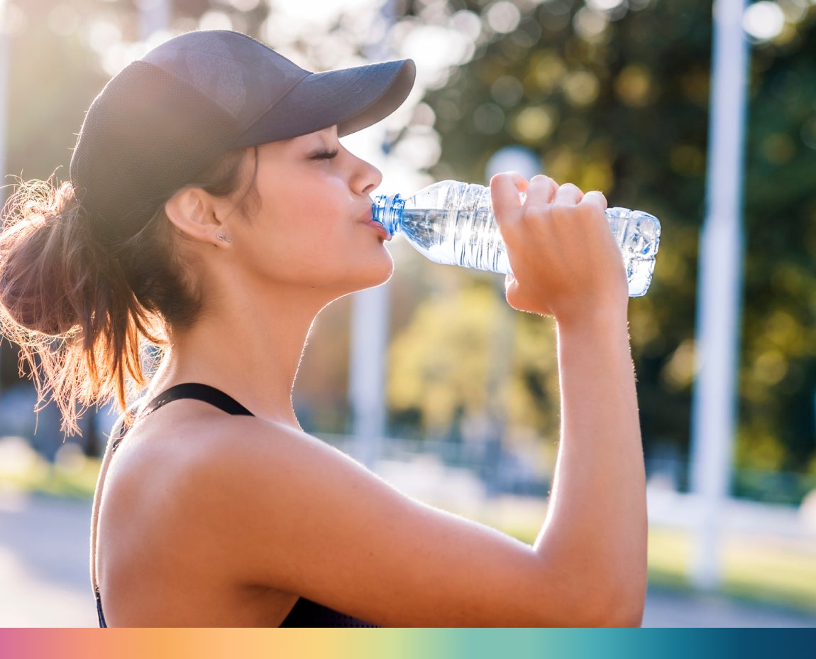 A woman wearing a baseball cap and athletic wear drinks from a water bottle to keep herself hydrated, she is standing outdoors with trees in the background.